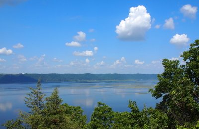 [View of the river almost looks like a large lake with greenery in the foreground, puffy clouds reflected as white dots on the water and the bluffs across the river providing a near-mountainous backdrop.]